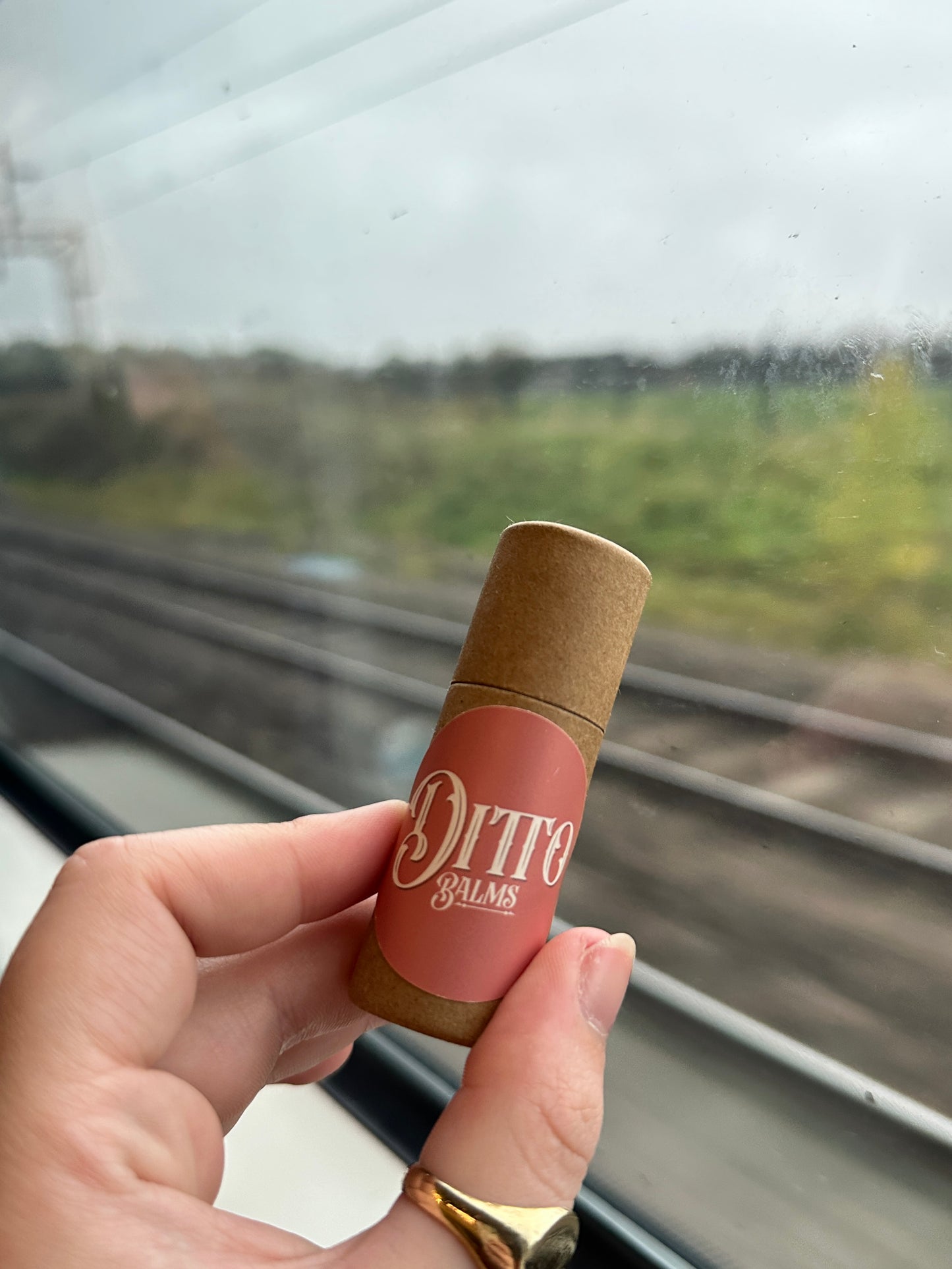 Hand holding a Ditto Balms solid perfume in front of a blurred train-track background