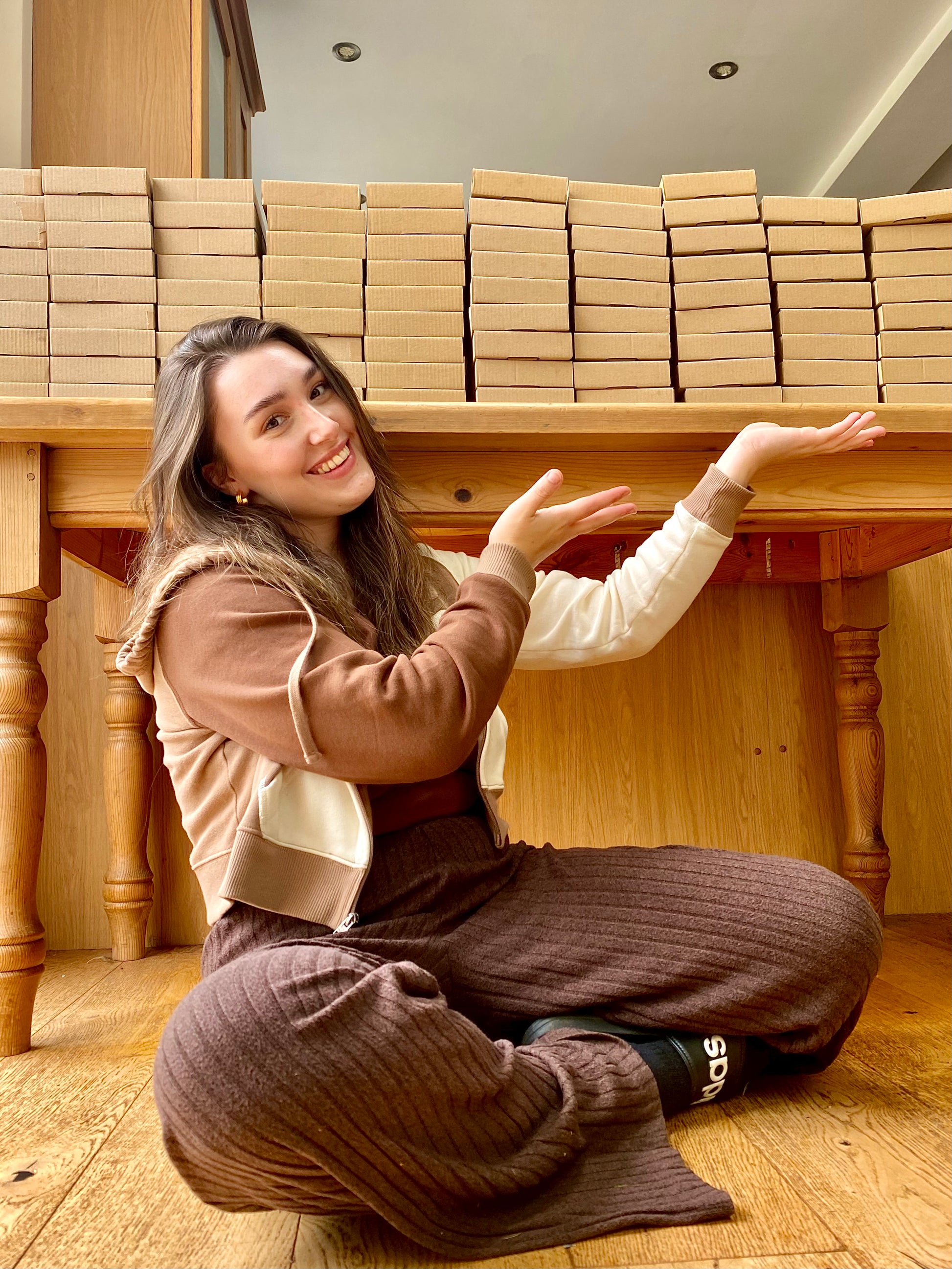 Emma sitting on the floor in front of a large stack of Ditto Bakes postal boxes in a room with wooden flooring.